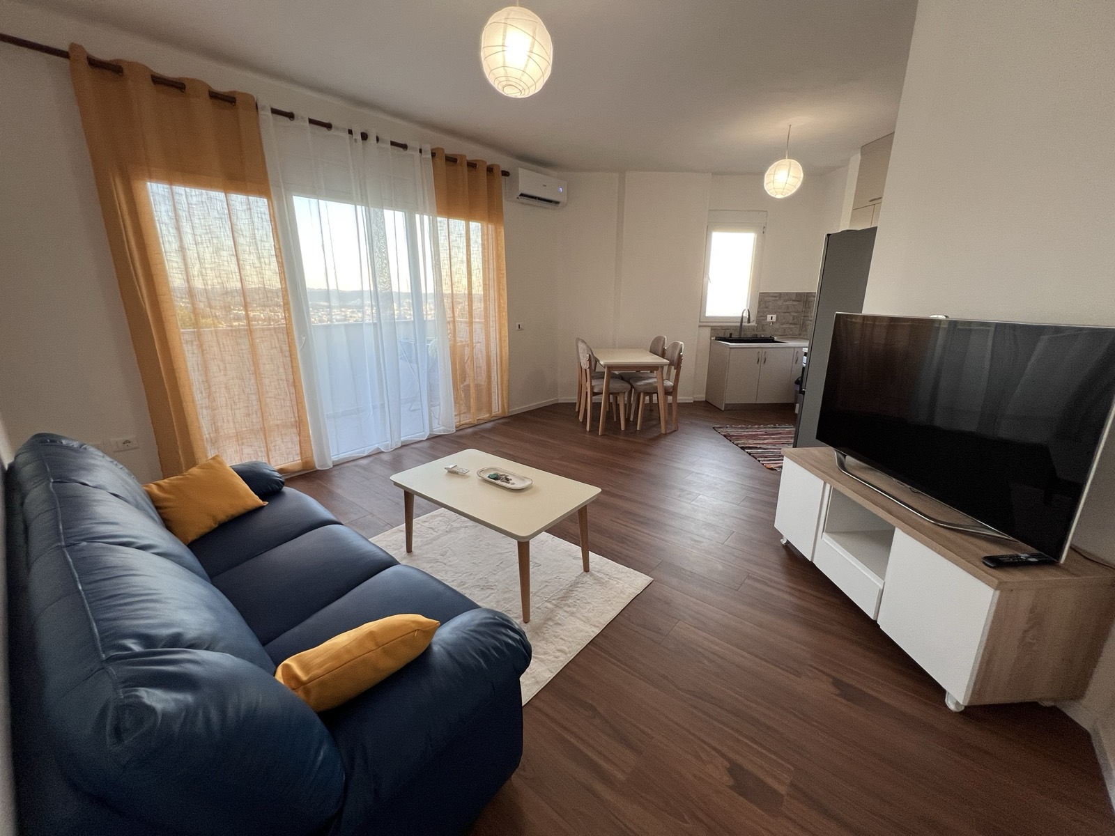 Open-plan living room with a blue sofa, dining table and floor-to-ceiling windows onto the balcony and city view